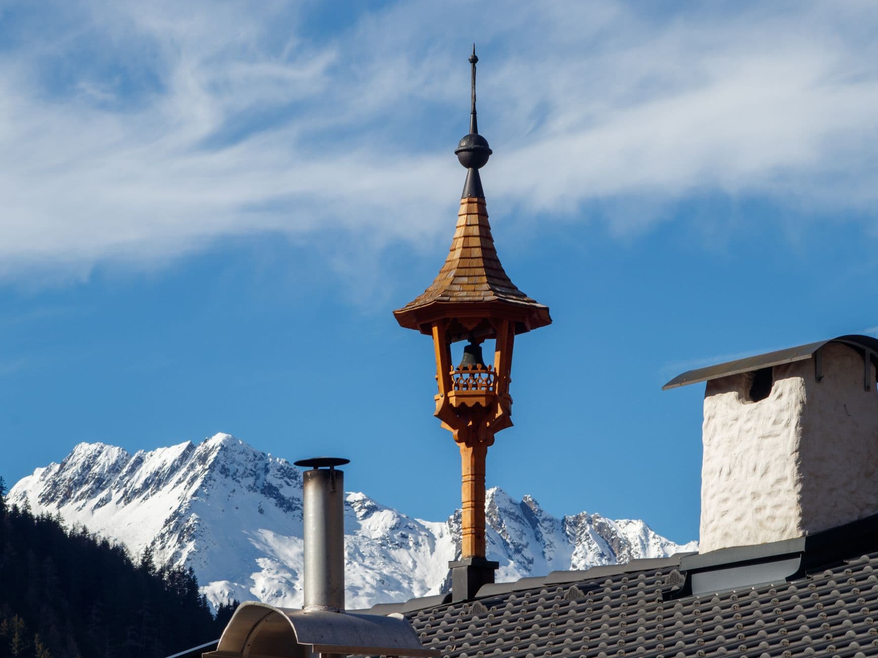 Alpine rooftop with ornate tower set against majestic snowy mountains under a clear blue sky.